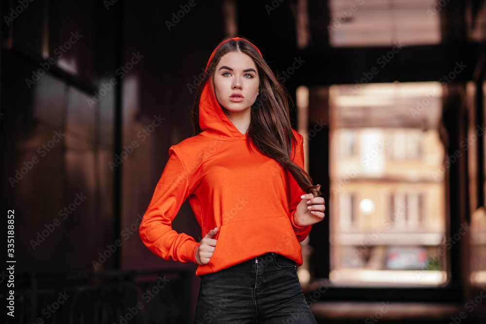 Portrait of glad brunette millennial female keeps both hands under chin, smiles pleasantly, wears orange bow headband, warm orange knitted sweater, expresses sincere feelings. Face expressions