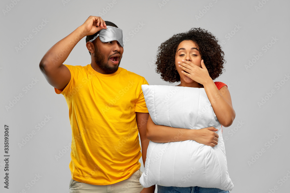 Obraz premium morning, bedtime and people concept - african american couple with eye sleeping mask and pillow yawning over grey background