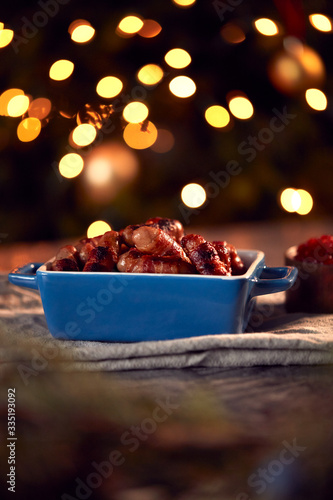 Traditional Dish Of Christmas Pigs In Blankets On Table Set For Meal With Tree Lights In Background © Monkey Business
