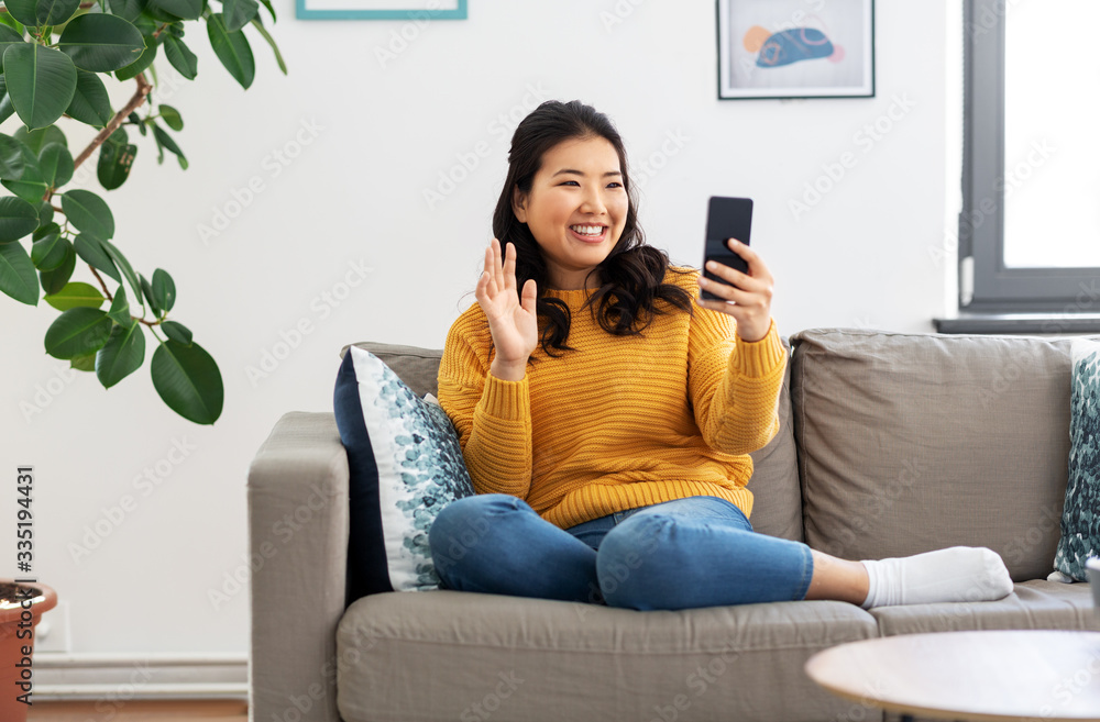 people, technology and communication concept - happy smiling asian young woman in yellow sweater sitting on sofa and having video call on smartphone at home people, technology and communication concept - happy smiling asian young woman in yellow sweater sitting on sofa and having video call on smartphone at home