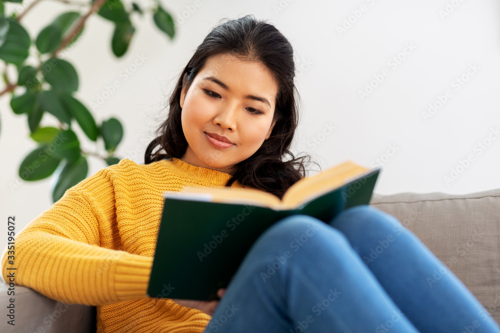 people and leisure concept - happy asian young woman in yellow sweater sitting on sofa and reading book at home people and leisure concept - happy asian young woman in yellow sweater sitting on sofa and reading book at home