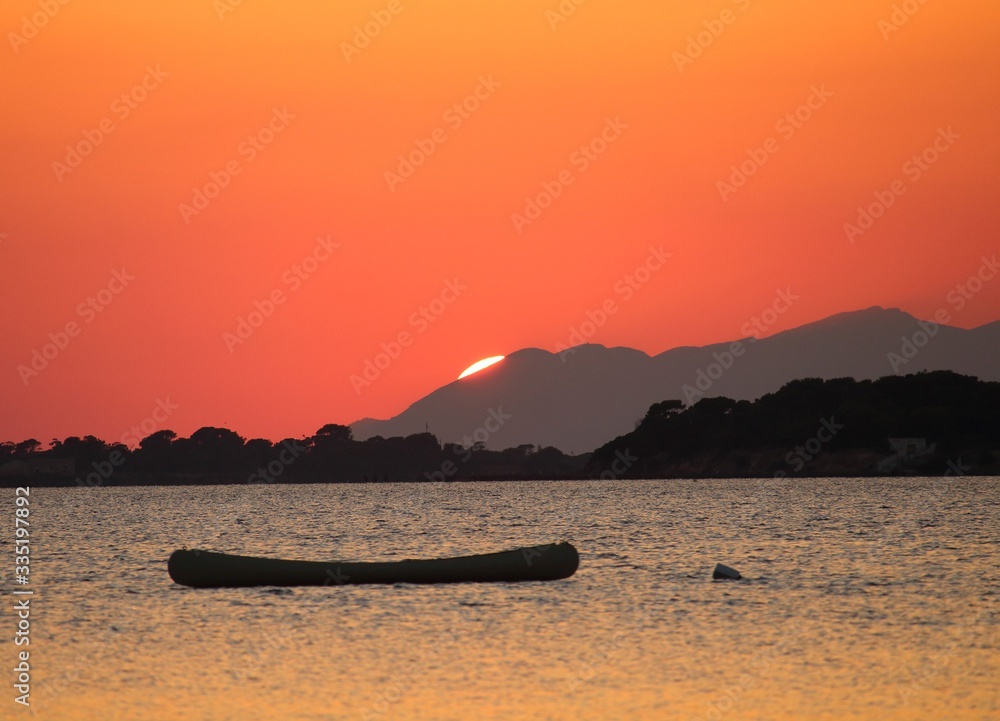 foreground of canoe moored in the sea with the sun setting behind the promontory