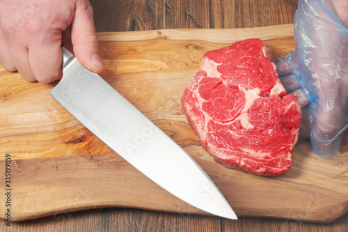 Rib eye steak on wooden cutting board, professional butcher with knife in his hand, Butcher dressed in white coat, red apron and blue glove.