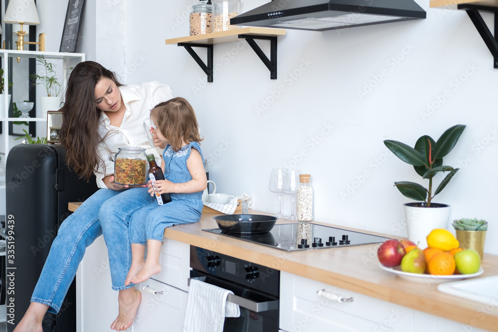 happy mom and daughter are cooking in the kitchen. mom brunette in white shirt and jeans, daughter in denim overalls. bright and spacious kitchen in Scandinavian style. a plate of fruit on the table happy mom and daughter are cooking in the kitchen. mom brunette in white shirt and jeans, daughter in denim overalls. bright and spacious kitchen in Scandinavian style. a plate of fruit on the table