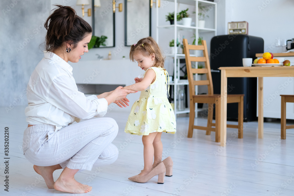 Beautiful mother and little daughter have fun in a bright and spacious apartment. The baby is trying on her mother's high-heeled shoes. Mom is a young brunette in white clothes, a girl in a dress. Beautiful mother and little daughter have fun in a bright and spacious apartment. The baby is trying on her mother's high-heeled shoes. Mom is a young brunette in white clothes, a girl in a dress.