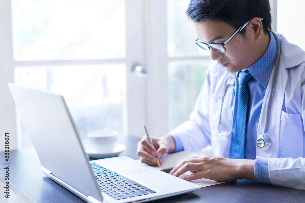 Young asian handsome doctor writing on notebook while using his laptop computer with serious face Young asian handsome doctor writing on notebook while using his laptop computer with serious face