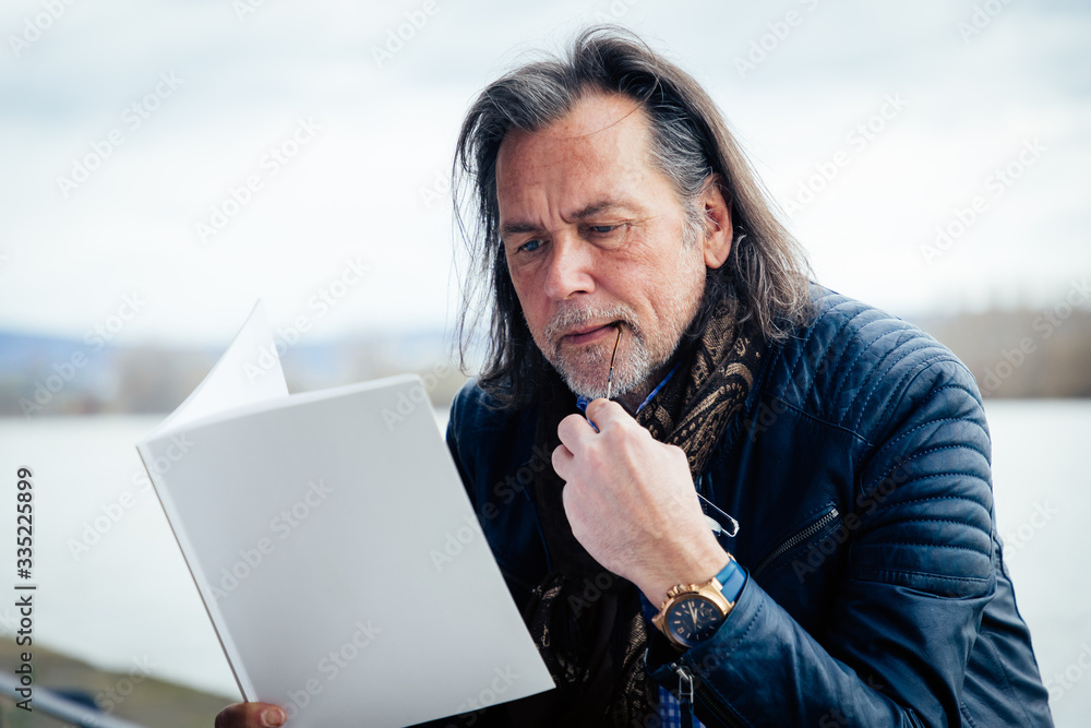 An elderly man with a beard and long grey hair sits on the bank of a river and reads a magazine with a blank cover Stock Photo | Adobe Stock An elderly man with a beard and long grey hair sits on the bank of a river and reads a magazine with a blank cover Stock Photo | Adobe Stock