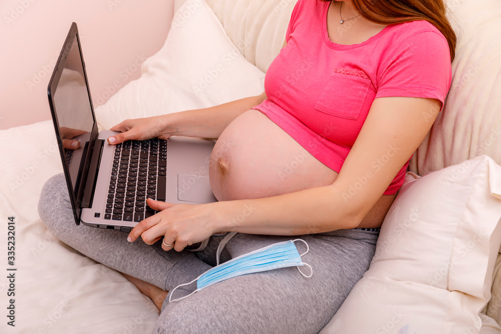 Naklejka premium Beautiful pregnant young woman working on a laptop sitting on the bed in the bedroom. Concept of female business, shopping, home office, quarantine, coronavirus covid 19 ncov. Stay at home concept.