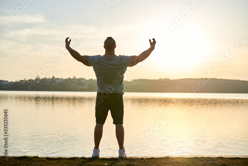Full-length portrait of young sportsman, wearing grey t-shirt, posing in front of lake at sunset. Muscleman poses, showing muscles, looking at the sun. © Natalia