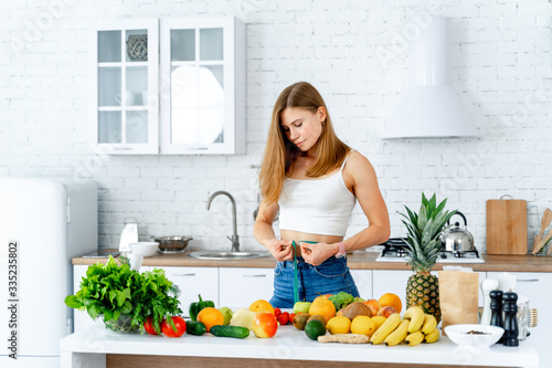 Young slim girl with measuring tape. Womn standing in the modern kitchen. Fruits and vegetables on the table. Fitness woman and healthy food concept.