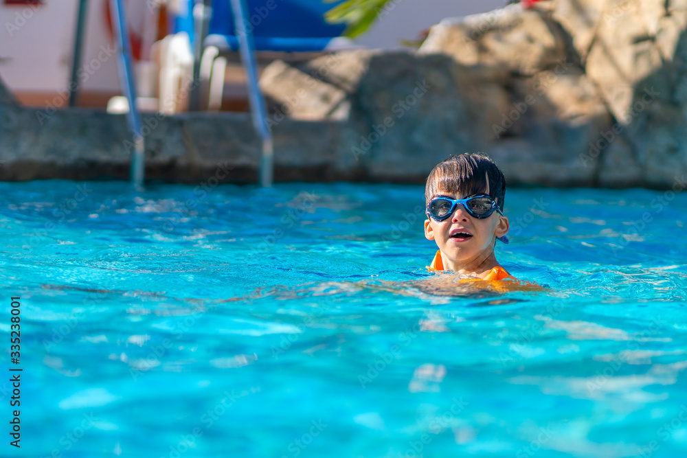 A happy boy in swimming goggles and arm ruffles in an outdoor pool. The child learns to swim. Family holidays at a tropical resort.