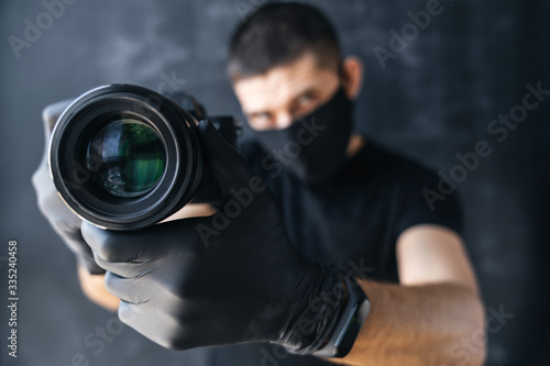 Photographer man in a medical black mask and gloves holds a camera photographs on a dark background. Photo business, epidemic and pandemic coronovirus.