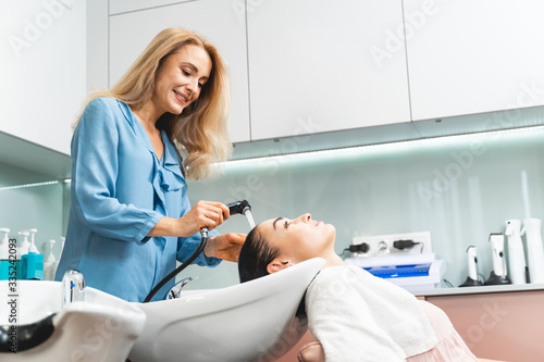 Positive delighted blonde hairdresser washing hair in sink