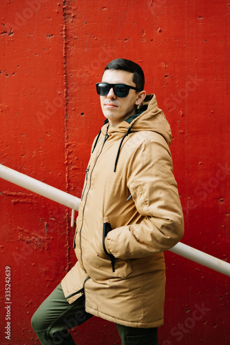 a young guy in a brown jacket and glasses on the street, standing next to a red wall on the stairs. street style