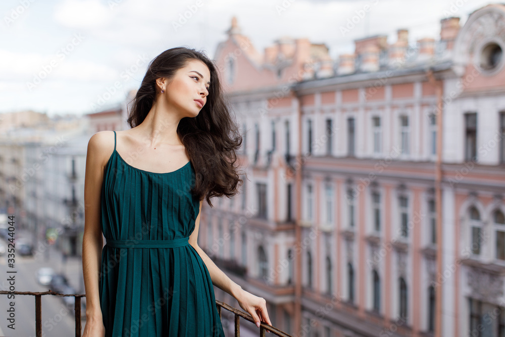Girl with long black hair in a green dress stands on a city balcony