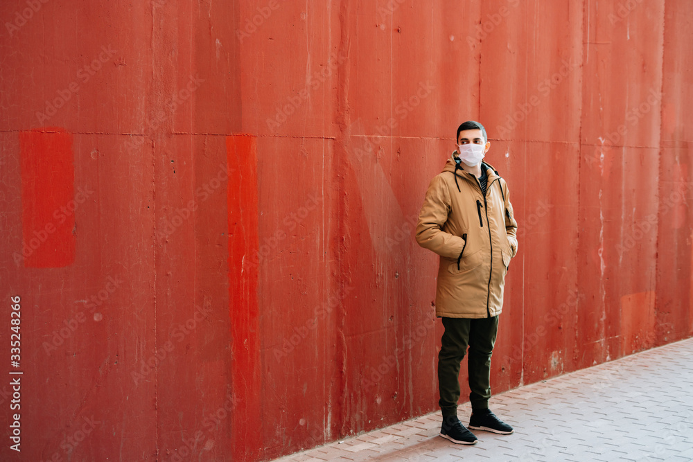 Obraz premium a young man in a brown jacket with a medical mask on his face, fighting against the corona 19 virus infection on the street, near the red wall.