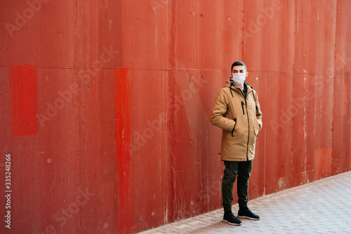 a young man in a brown jacket with a medical mask on his face, fighting against the corona 19 virus infection on the street, near the red wall.