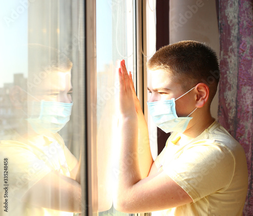 Portrait of a Caucasian boy in a medical mask stands and looking out the window. Misses you at home. Quarantine protection against coronavirus. Home insulation