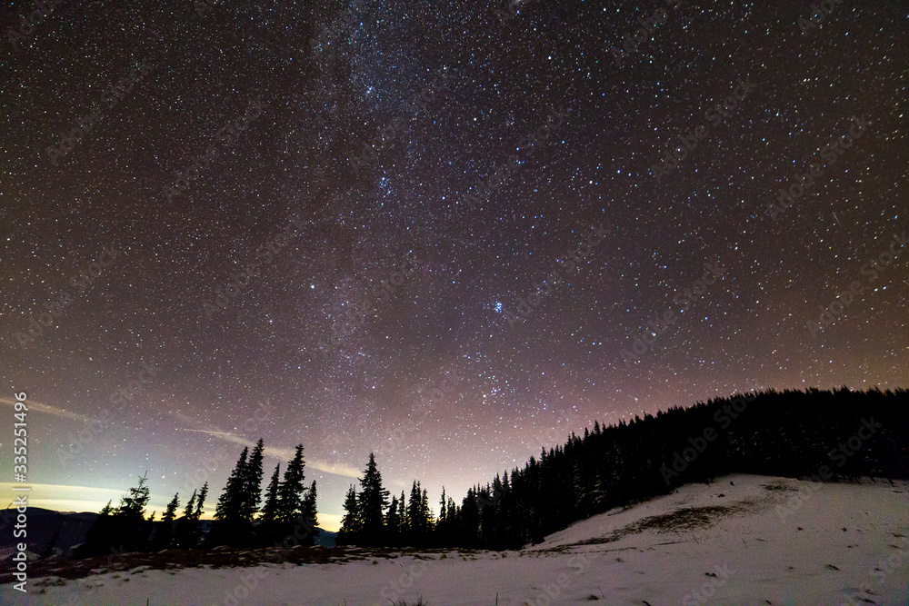 Winter mountains night landscape panorama. Milky Way bright constellation in dark blue starry sky over dark spruce pine trees forest, soft glow on horizon after sunset. Wide angle shot.