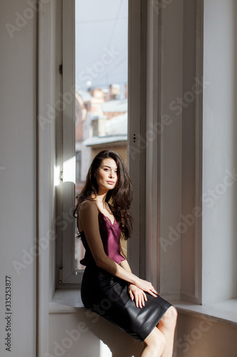 Brunette girl in a leather skirt and a wine blouse sits near a window on a windowsill in the sun