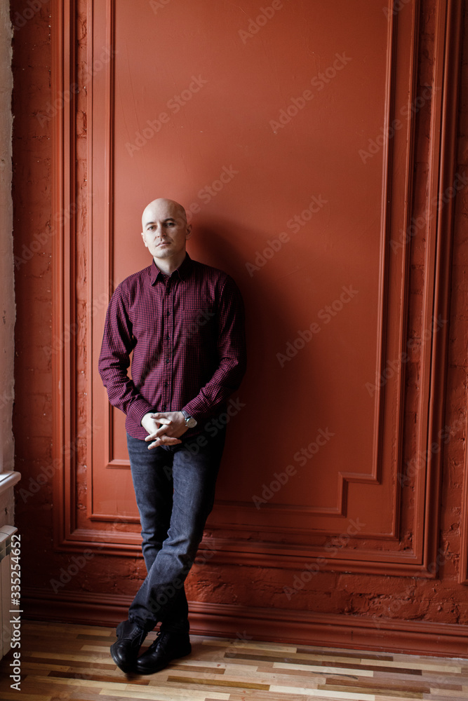 Bald man in a plaid shirt and jersey jeans stands against a background of an orange wall Bald man in a plaid shirt and jersey jeans stands against a background of an orange wall