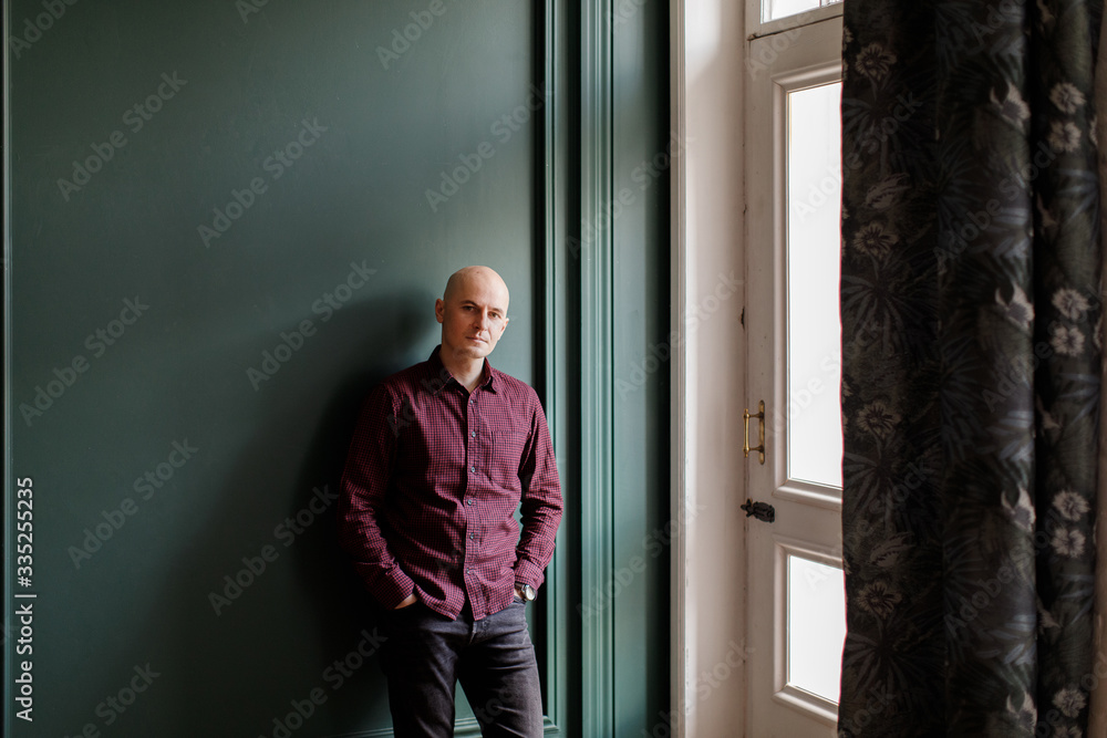 Bald man in a plaid shirt and gray jeans stands near a window against a green wall Bald man in a plaid shirt and gray jeans stands near a window against a green wall