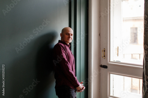 Bald man in a plaid shirt and gray jeans stands near a window against a green wall