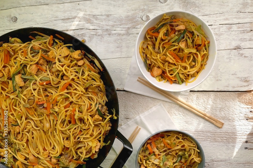 Stir-fry noodles with chicken, vegetables and portobello mushrooms. Wok and two bowls with chopsticks on a rustic wooden table. Top view.