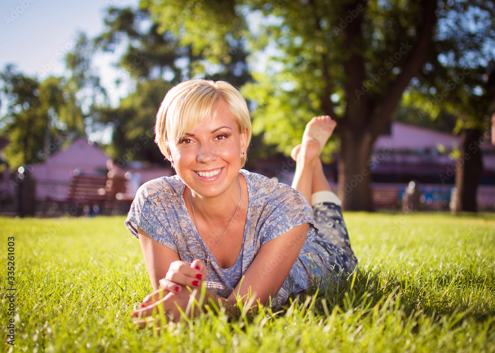 A smiling blonde girl with a short haircut is lying in a city park on green grass. A woman is resting in a park and looking into the camera A smiling blonde girl with a short haircut is lying in a city park on green grass. A woman is resting in a park and looking into the camera