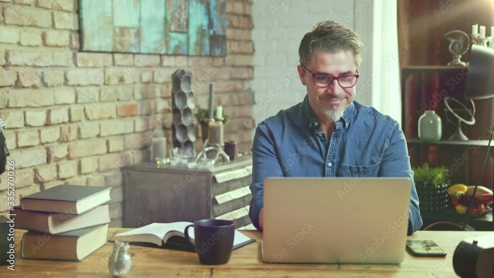 Older man tele working with laptop computer at home office in living room, thinking, looking busy.