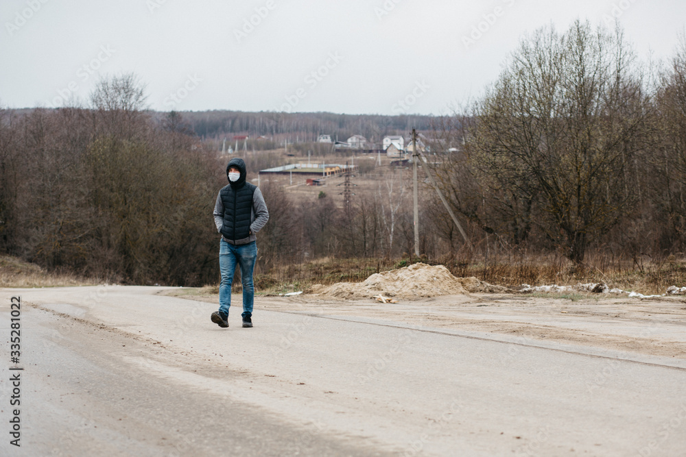 Obraz premium A man in a protective medical mask - modern measures of protection against coronovirus in quarantine conditions. A young man in casual style walks along an empty road