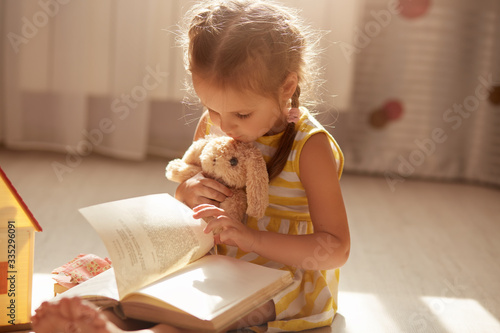 Quadro em tela Close up portrait of cute toddler girl with dark hair reading book for her soft toy while sitting on floor in her bedroom, reads fairytale to her rabbit
