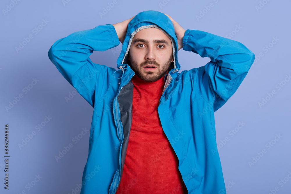 Close up portrait of young bearded man in hood with a bit opened mouth, looking horrified and shocked because of terrible mistake, keeps hands on head, looks at camerra, posing against blue background Close up portrait of young bearded man in hood with a bit opened mouth, looking horrified and shocked because of terrible mistake, keeps hands on head, looks at camerra, posing against blue background