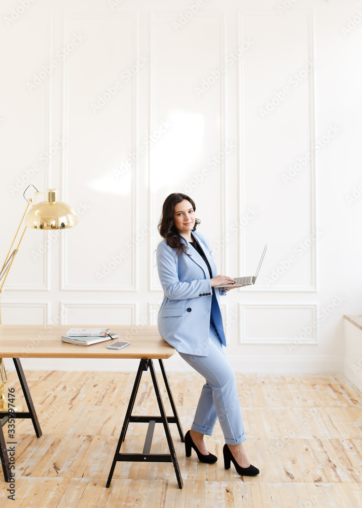 A serious business working freelance girl a woman with black hair in a pale blue suit working on a laptop and phone at a wooden Desk against a white wall in the home office A serious business working freelance girl a woman with black hair in a pale blue suit working on a laptop and phone at a wooden Desk against a white wall in the home office