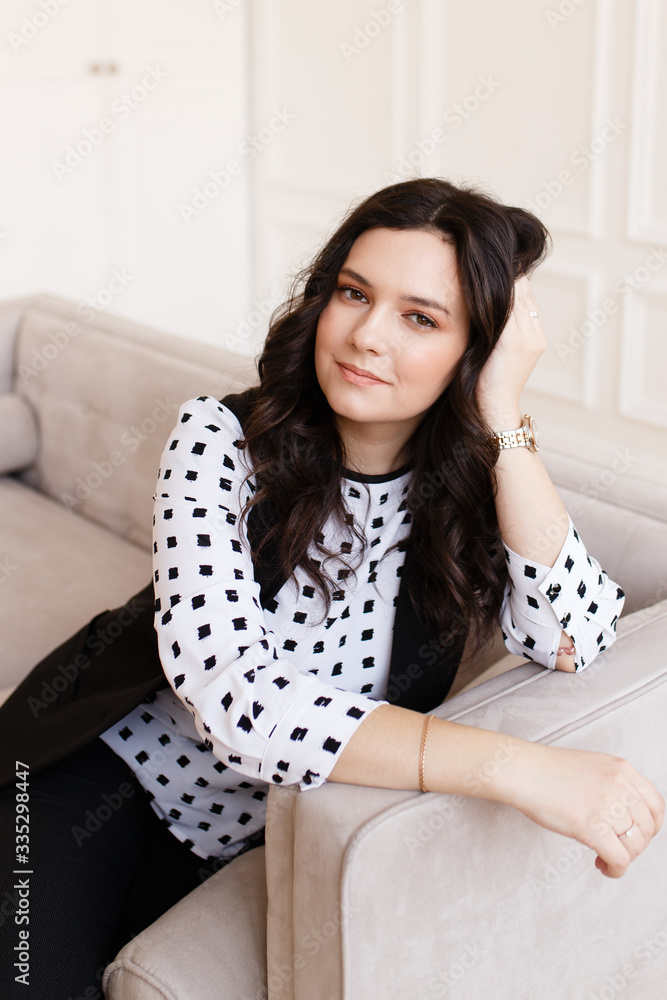 Business portrait of a serious working pretty girl freelancer businesswoman with black hair in a stylish black suit and a polka dot blouse sitting on a gray sofa at home on a white background Business portrait of a serious working pretty girl freelancer businesswoman with black hair in a stylish black suit and a polka dot blouse sitting on a gray sofa at home on a white background