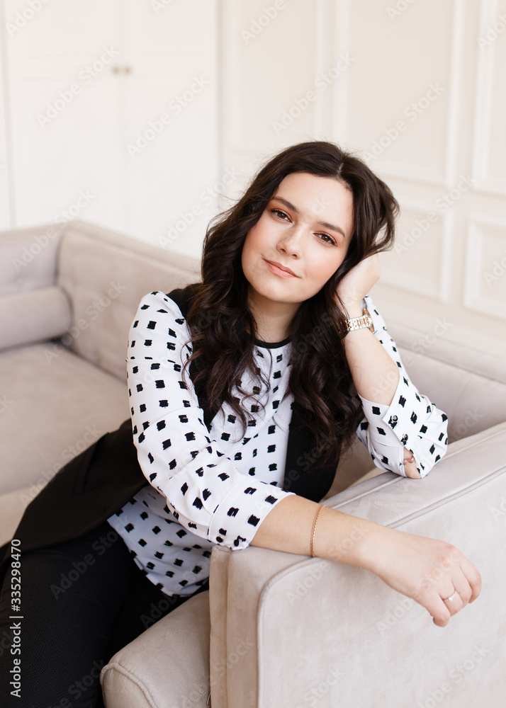 Business portrait of a serious working pretty girl freelancer businesswoman with black hair in a stylish black suit and a polka dot blouse sitting on a gray sofa at home on a white background Business portrait of a serious working pretty girl freelancer businesswoman with black hair in a stylish black suit and a polka dot blouse sitting on a gray sofa at home on a white background