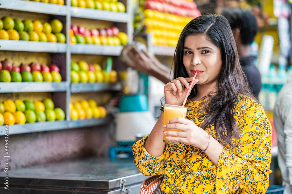 Young happy smiling Indian woman drinking orange juice outdoor in the market with colorful background of fruits. Young happy smiling Indian woman drinking orange juice outdoor in the market with colorful background of fruits.