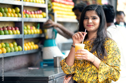 Young happy smiling Indian woman drinking orange juice outdoor in the market with colorful background of fruits.