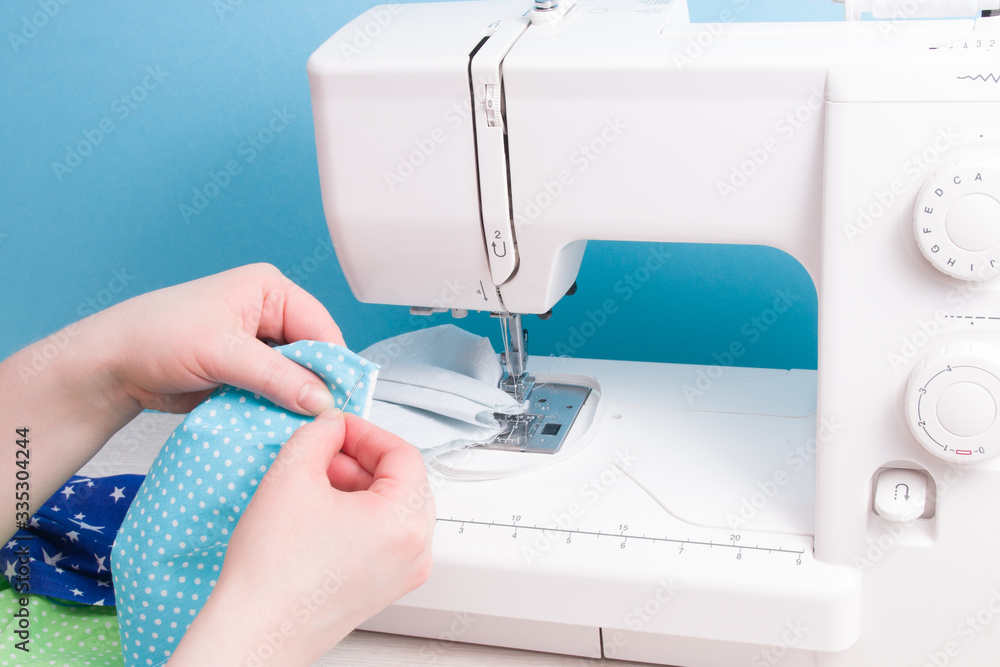 woman sews with a needle a protective face mask made of cotton fabric, hand sewing concept, sewing machine and mask on the background, blue background, virus protection concept, sewing process