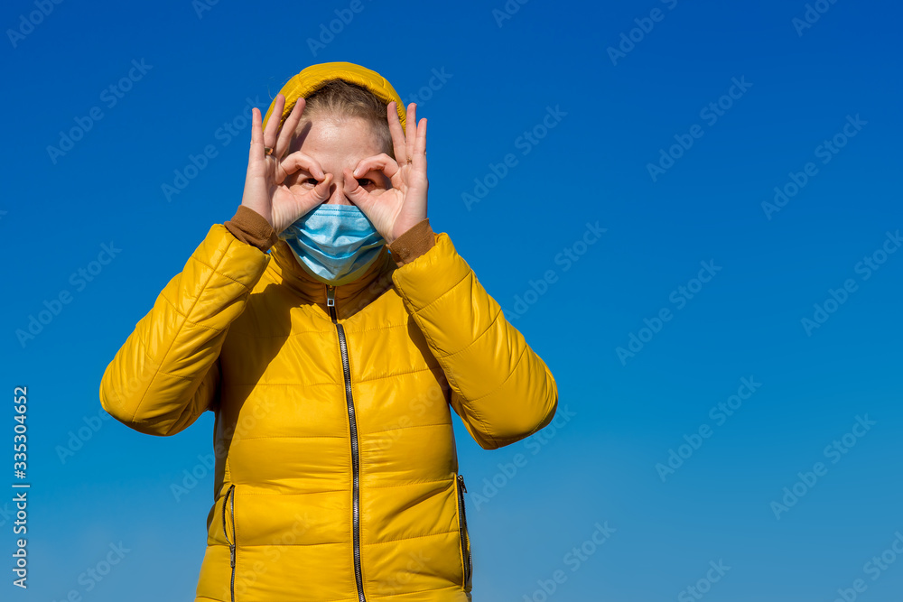 a girl in a yellow jacket with a hood and a blue medical mask holds hands in her face in the form of a batman mask, fun during quarantine. a girl in a yellow jacket with a hood and a blue medical mask holds hands in her face in the form of a batman mask, fun during quarantine.