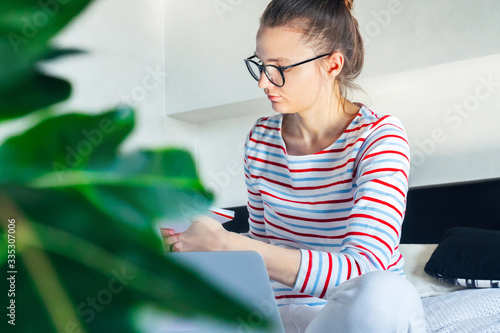young female student styding on distance education online on couch with laptop from home. freelancer work remotely, make write notes in online office, pay bills, invoice at quarantine self isolation. photo