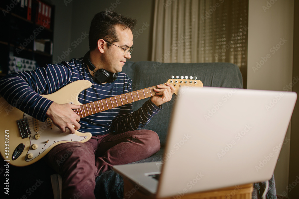 Obraz premium Boy taking an online course to learn to play the electric guitar. He is sitting on the sofa at home, playing the guitar in front of the laptop while listening to the lessons with headphones
