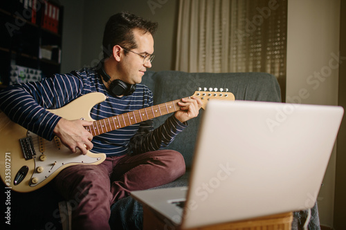 Boy taking an online course to learn to play the electric guitar. He is sitting on the sofa at home, playing the guitar in front of the laptop while listening to the lessons with headphones