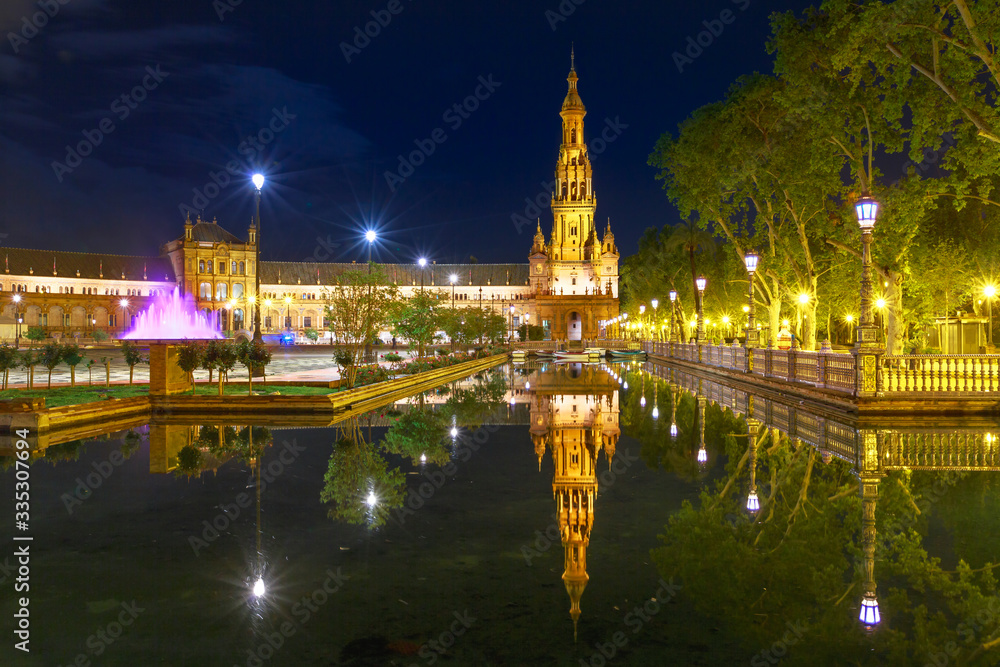Scenic panorama of Spain Square in Andalusia, Spain, illuminated at night. Renaissance building and one of two towers in Plaza de Espana reflects on canal of Guadalquivir river in Seville.