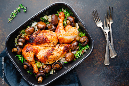 Top view of roasted chicken legs and breasts with fresh salad and mushrooms in black dish.