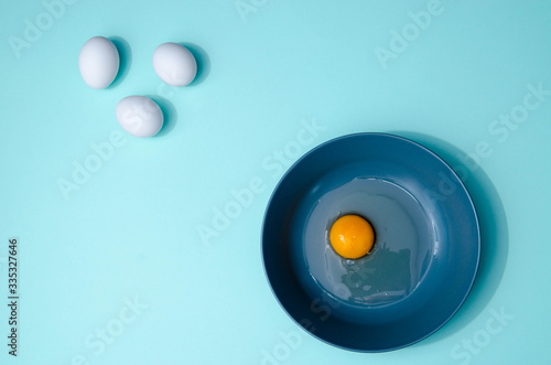 Three white eggs and a blue plate with broken egg on the table on the mint blue background