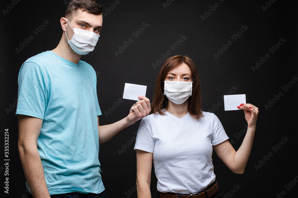 Studio stock portrait of two young caucasian people in aseptic masks holding two blank cards with place for text. Isolate on black.