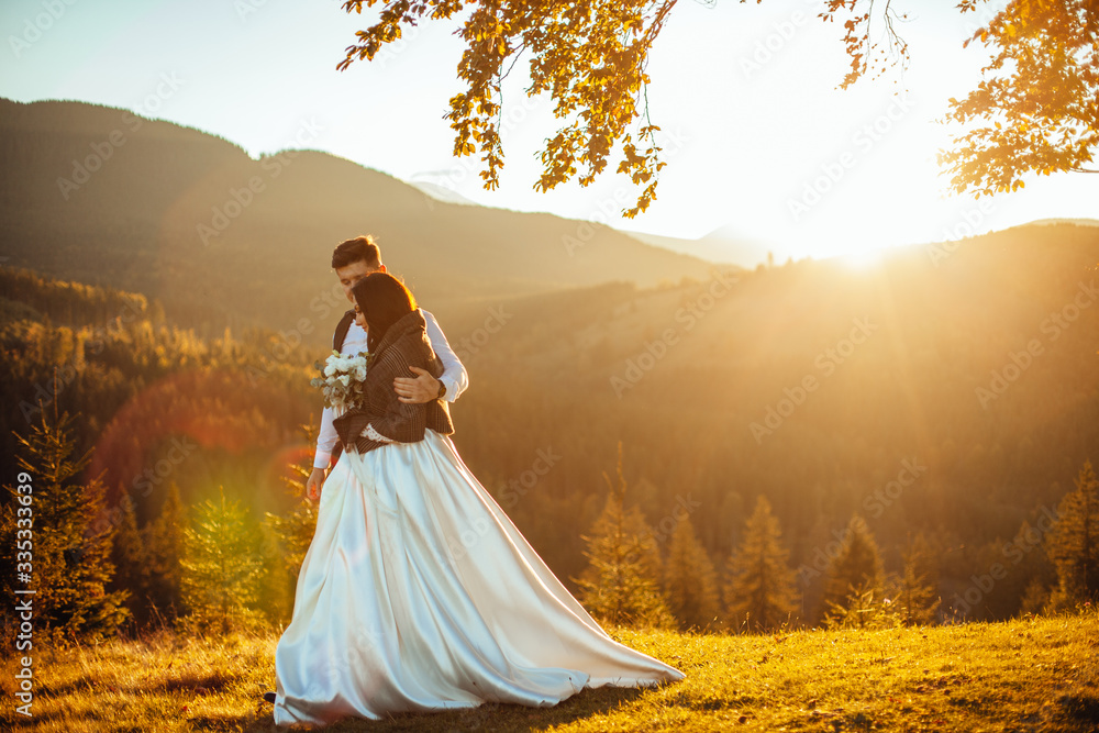 Beautiful young wedding couple of groom and bride standing on the green slope, hill. Sunset in Carpathian mountains