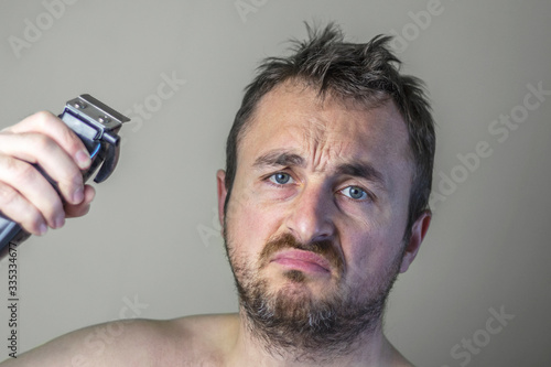 Portrait of a sad hairy man with unshaven face holding an electrical hair clipper.