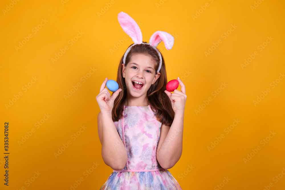 Studio portrait of smiling young girl wearing traditional bunny ears headband for easter. Brunette female with pigtails holding traditional colored eggs over yellow background. Close up, copy space. Studio portrait of smiling young girl wearing traditional bunny ears headband for easter. Brunette female with pigtails holding traditional colored eggs over yellow background. Close up, copy space.